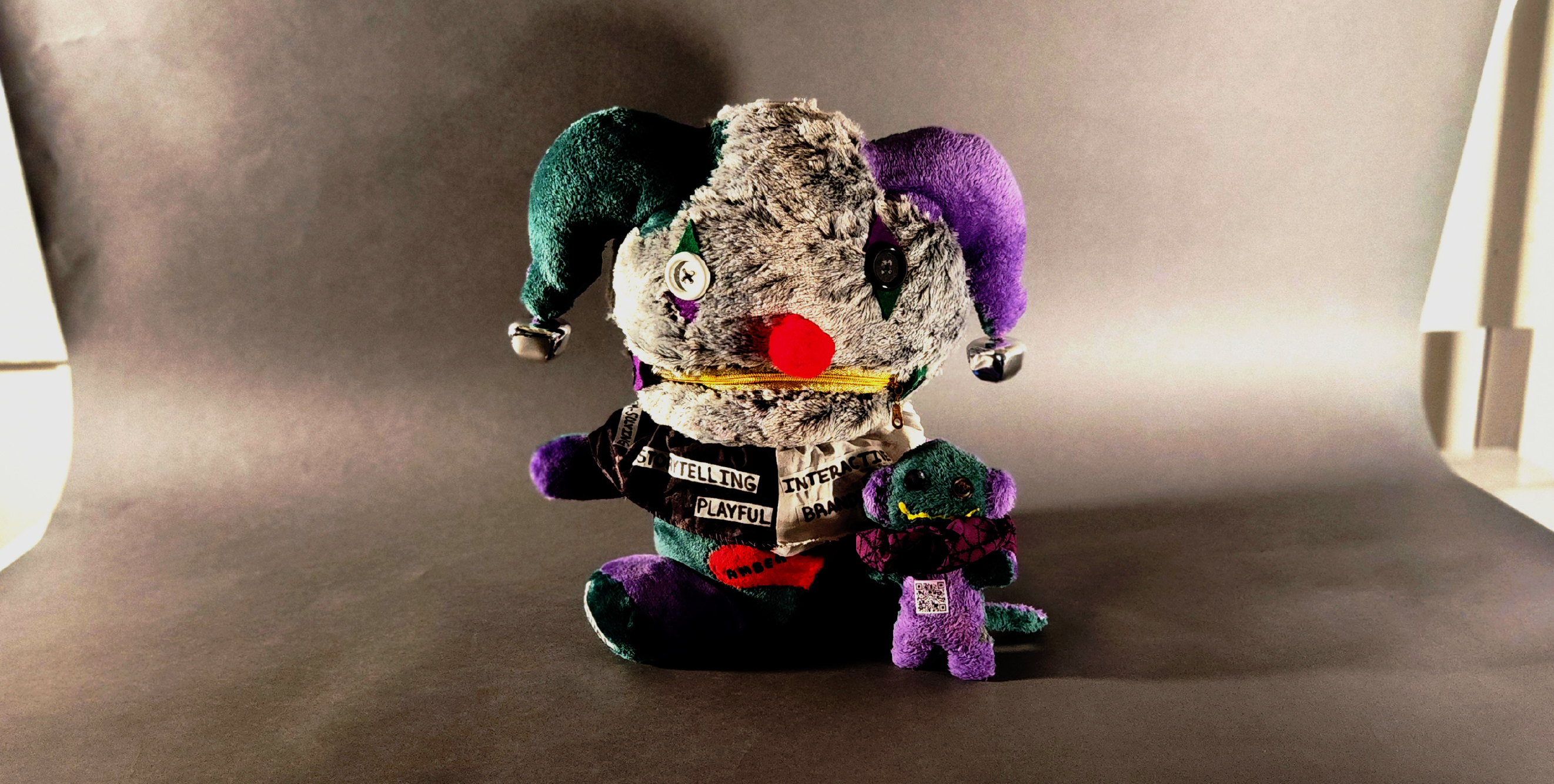 A beauty shot iamge of two plushies one bigger that looks like a jester ears with a clown red nose with colors gray, green and pruple and a yellow zipper mouth and black and white button eyes. On the right hadn side of the big plush is a tiny monkey like plush with a yellow smile and a QR code in the middle of its belly.