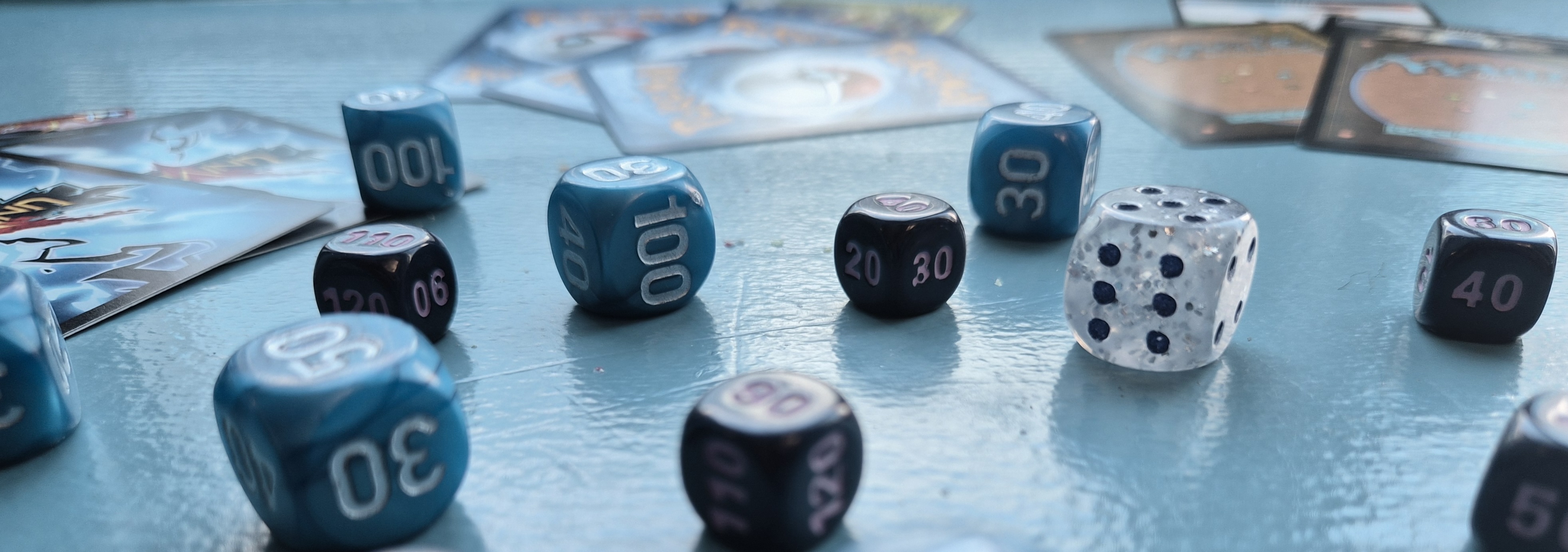 Photo image with different types of trading card games in the background that little out of view, center view dices of blue, purple, and a clear glitter dice in the center of image.
